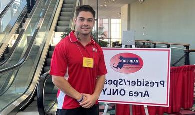 Gabe Elias stands in front of sign wearing a red shirt with hands clasped in front of him 